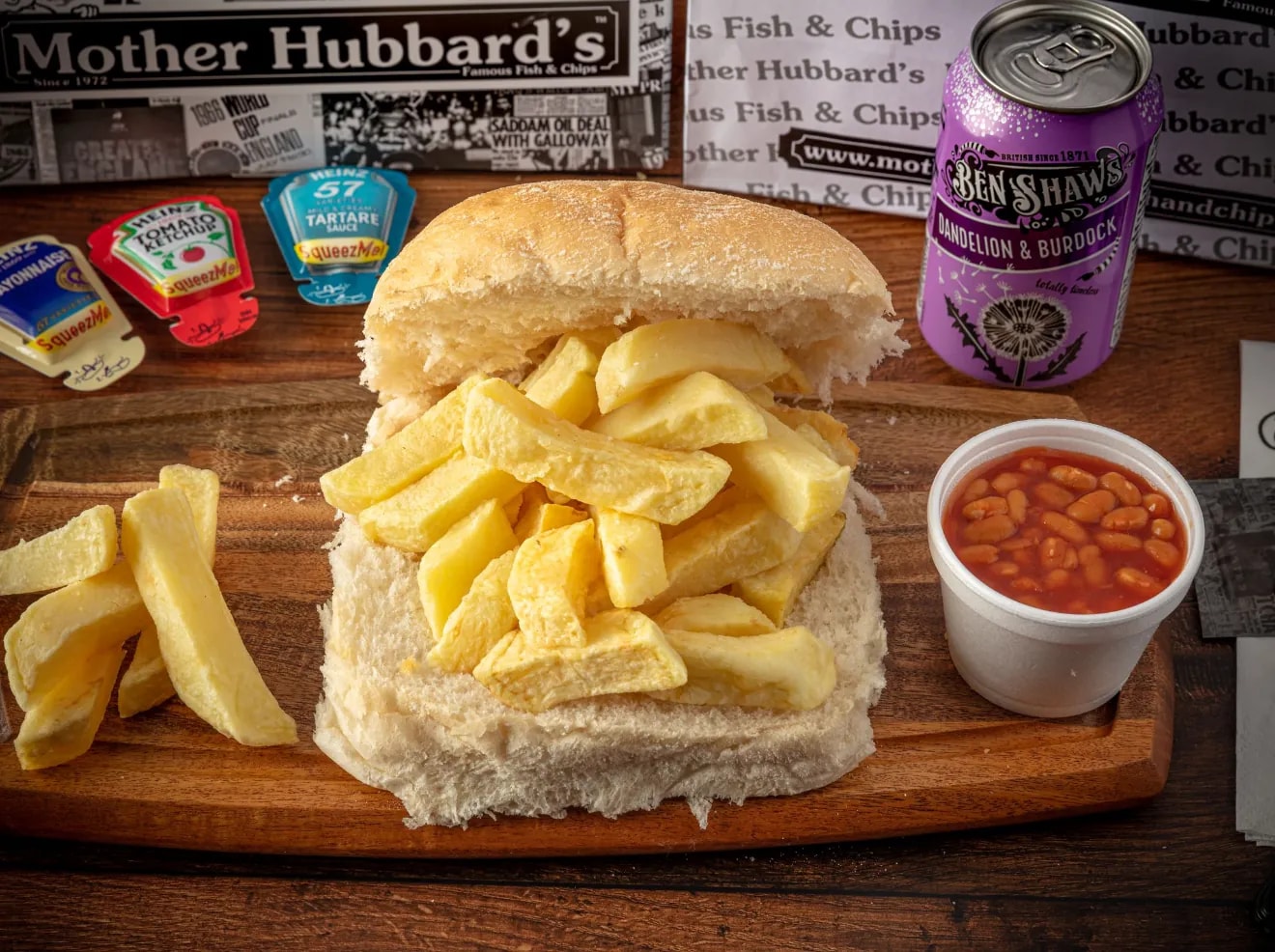 A chip butty: a soft bread roll filled with chips, beside baked beans, sauce sachets and a can of Ben Shaw’s
