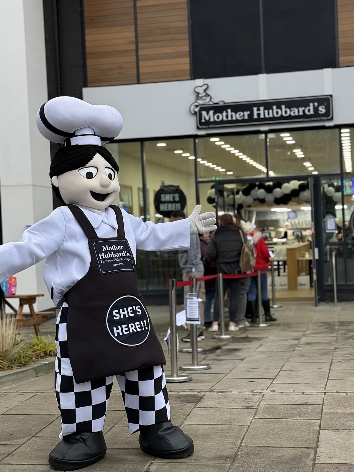 The Mother Hubbard's mascot in a chef's hat and apron welcoming a queue at a new shop's opening